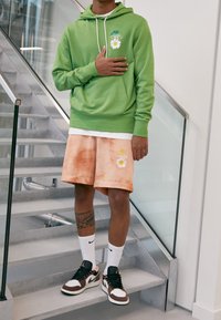 Zelená mikina s květinovým potiskem, oranžové šortky s technikou tie-dye, bílé ponožky a černé/hnědé tenisky. Na noze je viditelné tetování.