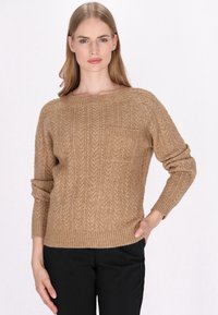 Beige kötött pulóver, bordázott textúrával, kerek nyakkivágással és egy kis zsebével a bal mellen. Fekete nadrággal párosítva.