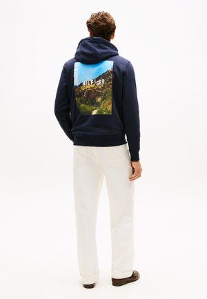 Άνδρας που φοράει μπλε ναυτικό φούτερ με εκτύπωση βουνού και κείμενο στην πλάτη, άσπρο παντελόνι και καφέ παπούτσια, στραμμένος με την πλάτη σε απλό φόντο.