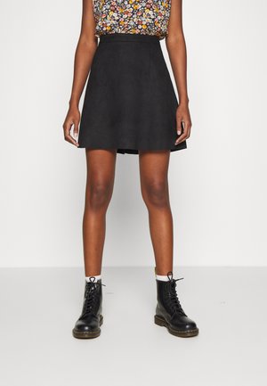 Kobieta ubrana w czarną spódnicę w linii A, kwiecisty bezrękawnik, białe skarpetki i czarne sznurowane buty wojskowe na jednolitym białym tle.