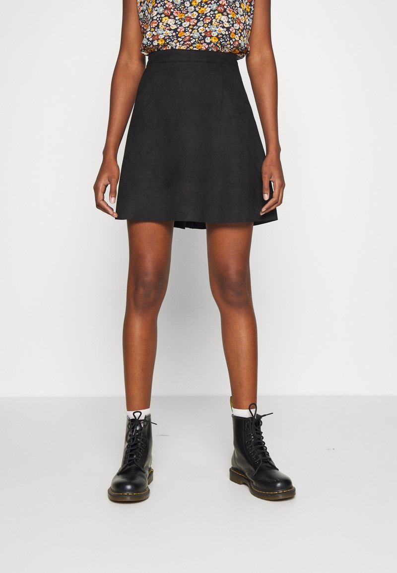 Femme portant une jupe noire trapèze, un haut sans manches à motifs floraux, des chaussettes blanches et des bottes de combat noires à lacets sur un arrière-plan blanc uni.