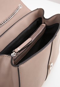 Béžová kožená kabelka s černým interiérem, obsahující zipovou kapsu a klopu na cvok. Elegantní řetězcový detail podél otvoru.