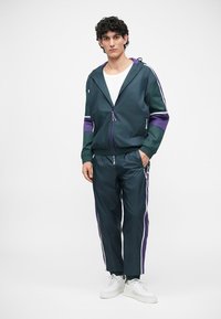 Pánsky športový teplákový set v tmavozelenej farbe s fialovými akcentmi, ktorý obsahuje zipsovú bundu a elastické nohavice so bočnými pruhmi a vreckami.