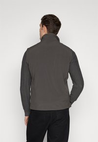 Gilet grigio imbottito con colletto alto e maniche a coste, che offre un design a strati. Texture liscia con modelli delle maniche a contrasto.