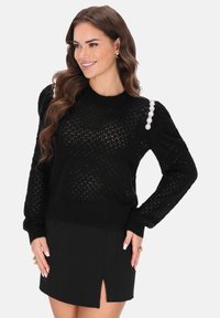 Fekete kötött pulóver mintás textúrával, gyöngy díszítéssel a vállon, egyszerű fekete szoknyával párosítva.