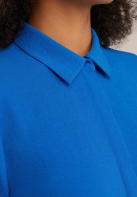 Primo piano di una persona che indossa una camicia colletto blu brillante con una patta dei bottoni nascosta e capelli ricci naturali.