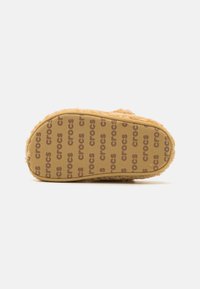 Zijaanzicht van een fuzzy, tan slip-on slipper met herhaaldelijk 'crocs' gedrukt op de rubberen zool tegen een witte achtergrond.