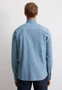 Chemise boutonnée à manches longues, bleu clair, avec une texture douce, coupe droite et ourlet arrondi. Elle possède des poignets avec des boutons et un col classique.