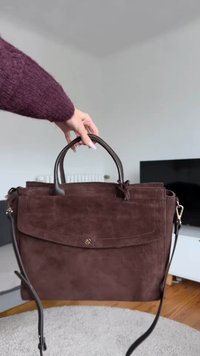 Brown suede handbag with dual handles, a flap closure, and adjustable shoulder strap. Features stitched detailing and a smooth texture.