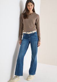 Femme portant un sweat à capuche marron côtelé, un jean large bleu avec une ceinture beige et des baskets beige, debout devant un mur blanc et beige.