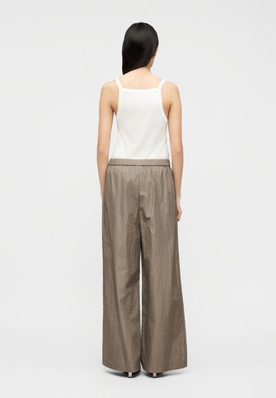 Γυναίκα που στέκεται από πίσω, φορώντας λευκή αμάνικη μπλούζα και φαρδιά, ψηλόμεσα παντελόνια σε αποχρώσεις ταουπέ, σε ουδέτερο φόντο.