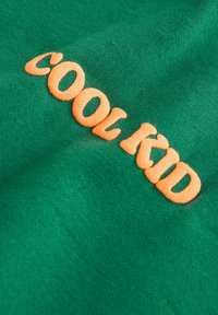 A zöld anyagon narancssárga dombornyomott hímzés látható, amely a "COOL KID" feliratot formázza. Az anyag puha tapintású, egységes felületű és enyhén fényes.