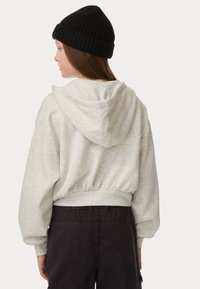 Lichtgrijze cropped hoodie met een trekkoordkap en ribgebreide manchetten, gecombineerd met zwarte broek. De stof lijkt zacht met een subtiele textuur.