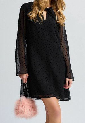 Femme portant une robe noire transparente à pois, tenant un sac à main rose duveteux avec une bandoulière chaîne, montrée des épaules jusqu'au milieu de la cuisse.
