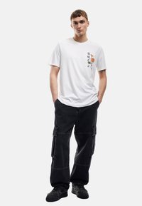 Camiseta de algodón blanca con bordado floral en el lado izquierdo del pecho, combinada con pantalones cargo negros de corte holgado que tienen bolsillos laterales.