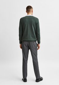 Uomo con maglione verde, camicia bianca, pantaloni grigi e scarpe eleganti nere in piedi di spalle contro uno sfondo bianco semplice.