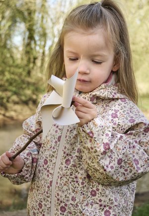Ragazza giovane con giacca floreale esamina un girandola di carta bianca attaccata a un bastoncino all'aperto, vicino a uno sfondo naturale sfocato.