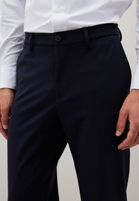 Mannelijke torsowent met een witte blouse met lange mouwen, ingestopt in zwarte formele broeken met een knoop en riemlussen, handen ontspannen aan de zijkanten.