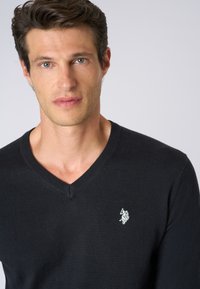 Maglione nero a scollo a V realizzato in materiale morbido, con un piccolo logo ricamato bianco sul lato sinistro del petto. Maniche corte, design aderente.