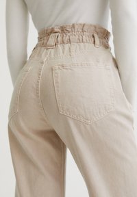 Beige, high-waisted denim broek met geplooide tailleband, twee achterzakken en een ontspannen pijpfit. Zachte textuur en zichtbare stikseldetails.