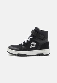 Baskets montantes noires avec semelle blanche, dotées d'un logo sur le côté, d'un laçage à l'avant et d'une sangle velcro ; fabriquées en cuir et en matériaux textiles.