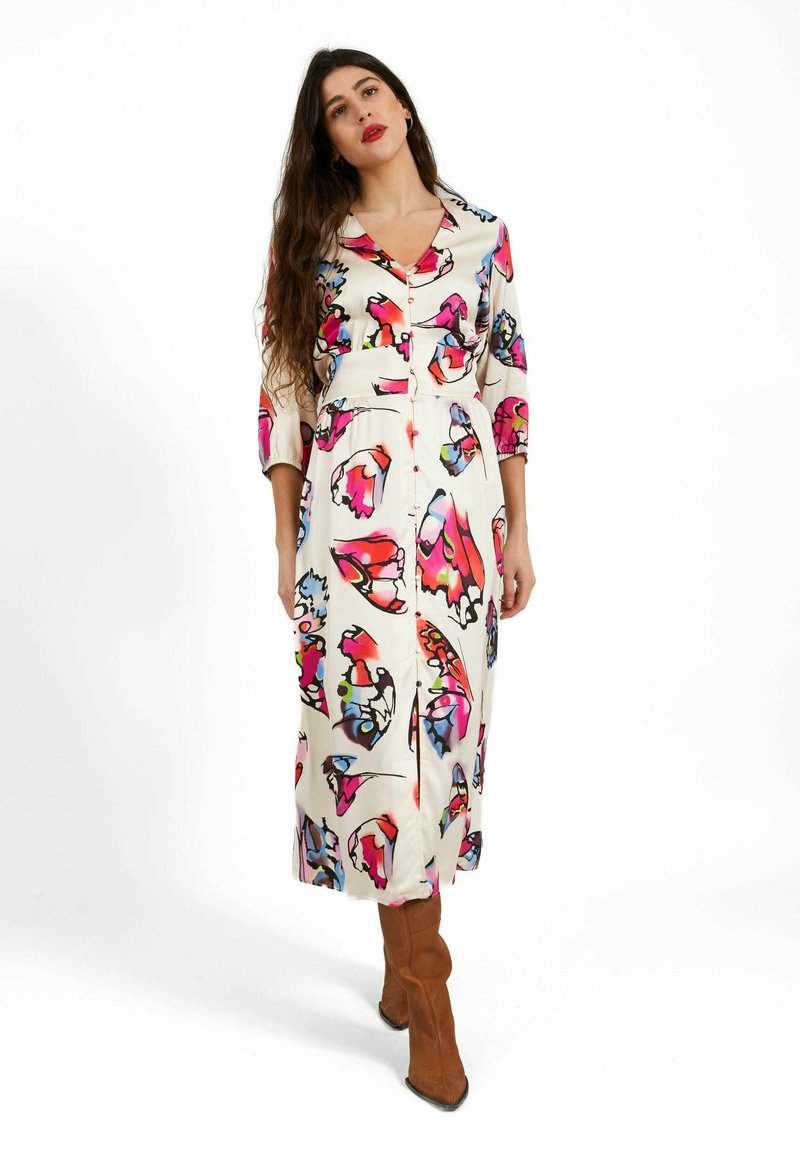 Femme aux longs cheveux foncés portant une robe midi blanche avec des imprimés colorés de papillons et des bottines marron à talons, debout sur un fond blanc.