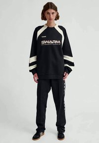 Felpa nera con accenti bianchi e logo "SWARM International". Abbinata a pantaloni neri con strisce bianche e sneakers nere.