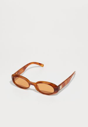 Brown tortoiseshell sunglasses with rounded lenses and thin, curved arms. Features light brown lenses and subtle metallic branding on the side.