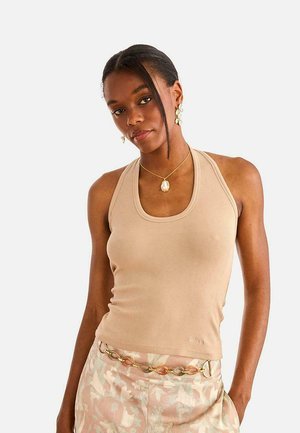 Mujer con top sin mangas beige, pantalones estampados con un cinturón de cadena y joyas de perlas sobre un fondo blanco.