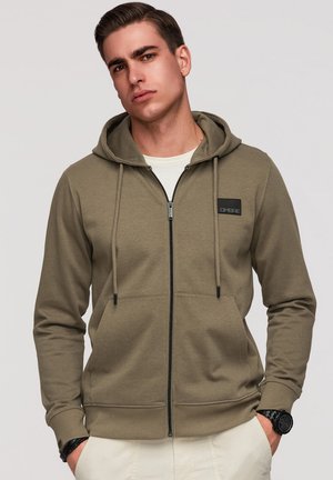 Jeune homme portant un sweat à capuche zippé vert olive avec cordons et une montre-bracelet noire, debout les mains dans les poches.