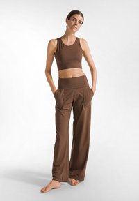 Donna in piedi a piedi nudi che indossa un top corto senza maniche marrone e pantaloni larghi abbinati con tasche su uno sfondo bianco.