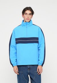 Ανοιχτό μπλε πούλεντζερ με ψηλό γιακά, ναυτικό ρίγα διαγωνίως στο στήθος, ελαστικούς καρπούς και φερμουάρ στο τέταρτο. Απαλή υφή και μινιμαλιστικός σχεδιασμός.