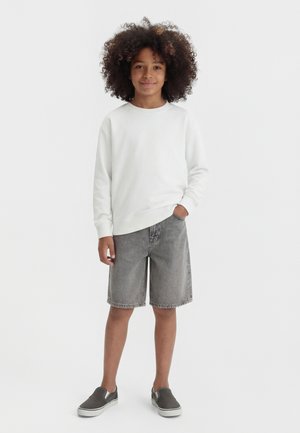 Παιδί με σγουρά μαλλιά που φοράει λευκό μακρυμάνικο πουκάμισο, γκρι τζιν σορτς και γκρι παπούτσια χωρίς κορδόνια, στέκεται με το ένα χέρι στην τσέπη.