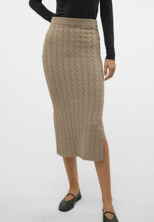 Vrouw in een beige kabelgebreide midi rok met een zijsplits, gecombineerd met een zwart lang mouwtop en zwarte platte schoenen.