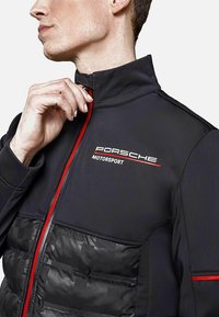 Czarna kurtka z pikowanej tekstury w dolnej części, z czerwonymi akcentami oraz białym logo "Porsche Motorsport" na piersi.