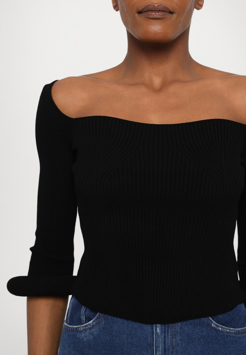 Black ribbed knit top with an off-shoulder design, three-quarter sleeves, and textured detailing, paired with blue denim jeans.