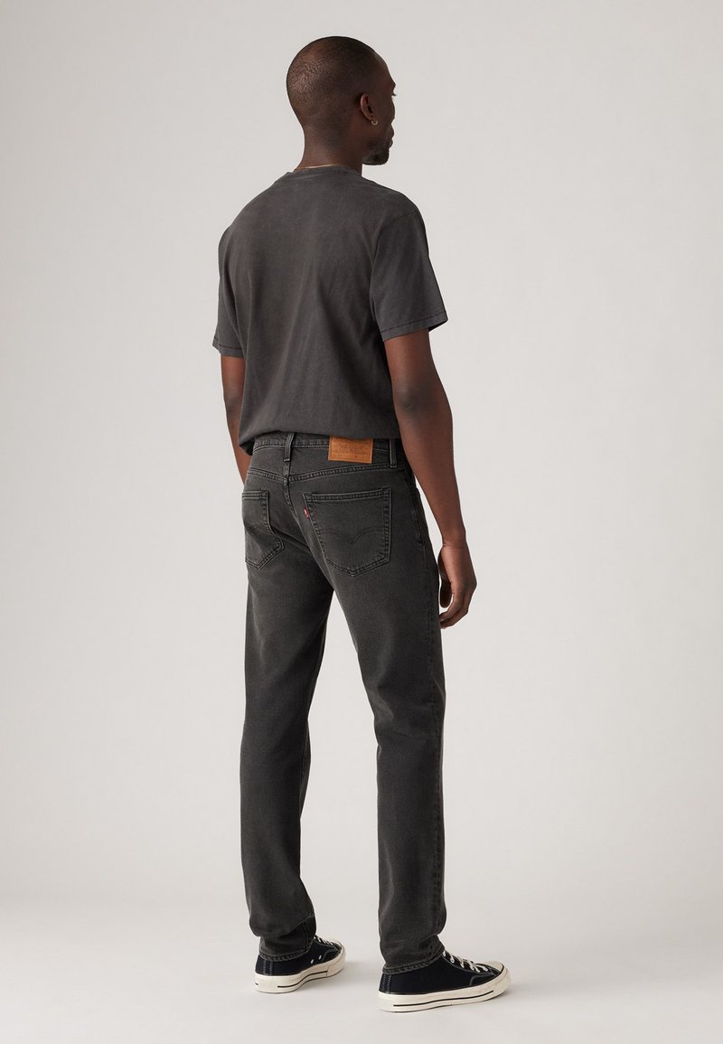Jeans de mezclilla negros con corte recto, presentan una etiqueta de cuero marrón en la parte trasera de la cintura, combinados con una camiseta gris oscura y zapatillas negras.