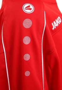 Rode sportshirt met een gestructureerde stof, witte zijaccenten en cirkelpatroon details in het wit nabij de schouder. Merklogo aanwezig.