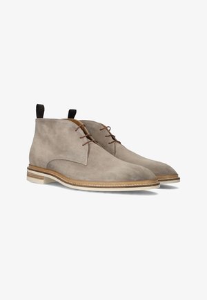 Beige Wildleder-Ankle Boots mit braunen Schnürsenkeln, einer runden Zehenpartie und einer weißen Sohle mit einem beige farbenen Akzentband sowie einer Schlaufe hinten.