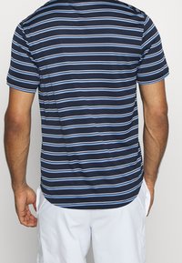 Muž v námořnickém modrém polo tričku s vodorovnými bílými a světle modrými pruhy a bílých šortkách, znázorněn zezadu.