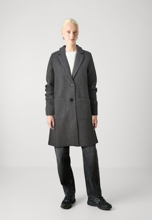 Manteau en laine gris avec un col cranté, fermeture à un bouton et deux poches à l'avant. Porté sur un haut blanc et associé à un pantalon sombre.