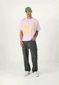 Jaunas vyras dėvi žalią kepurę, rožinį marškinėlį su geltonu abstrakčiu raštu, laisvas tamsiai pilkas džinsus ir baltus sportbačius, stovi su viena ranka kišenėje.