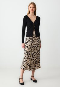 Zwarte geribde langwerpige cardigan met gouden knopen, gecombineerd met een beige rok met zwart zebrapatroon en zwarte platte schoenen met bandjes.