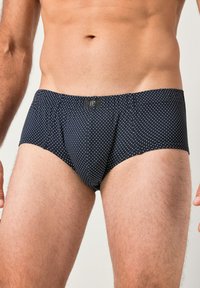 Torso maschile e parte superiore delle gambe che indossano slip blu navy con piccoli pois bianchi e una piccola etichetta nera al centro della fascia in vita.