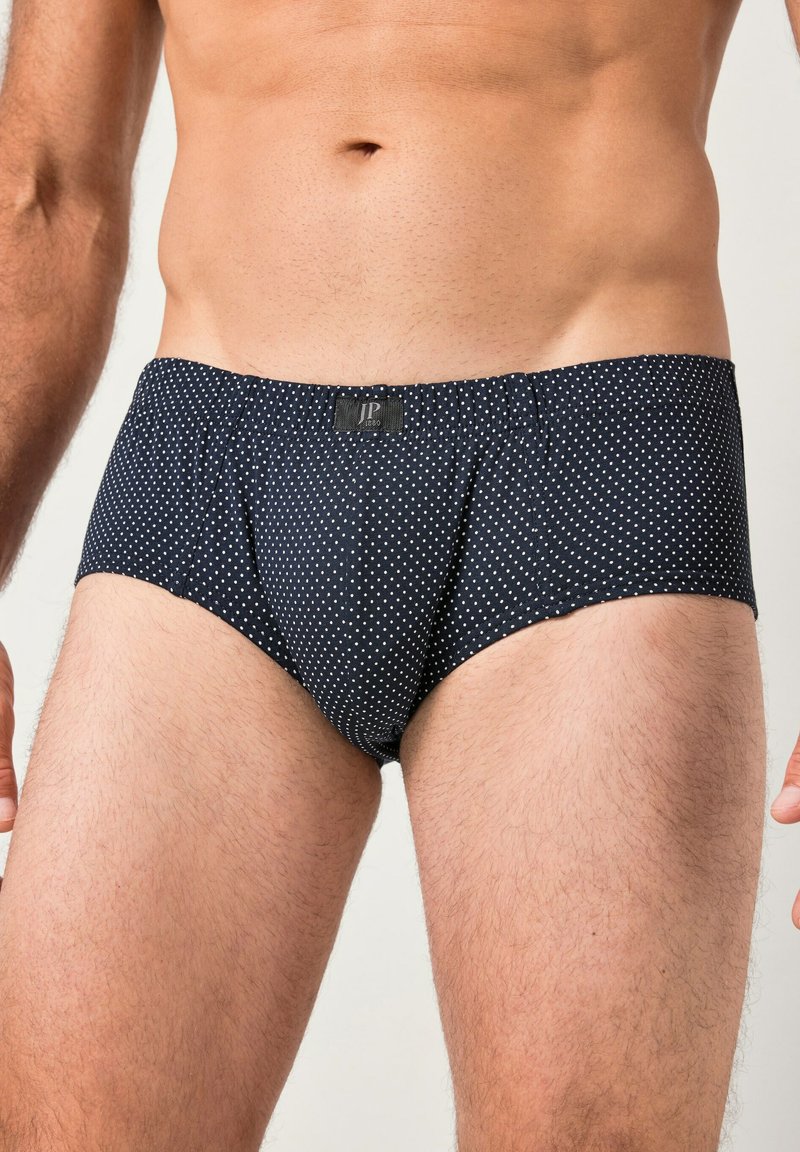 Torso maschile e parte superiore delle gambe che indossano slip blu navy con piccoli pois bianchi e una piccola etichetta nera al centro della fascia in vita.