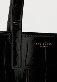 Svart skinnveske i lakkskinn med preget mønster, utstyrt med to solide håndtak og en gullfarget logo med teksten "TED BAKER LONDON." Jevn tekstur.