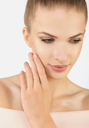 Gold ring with a minimalist design featuring a small gemstone and letter accent. Hand positioned near the face, showcasing clean lines.
