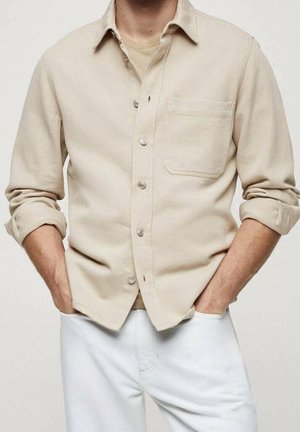 Homme portant une chemise beige boutonnée avec manches retroussées et un pantalon blanc, mains dans les poches, debout devant un fond uni.