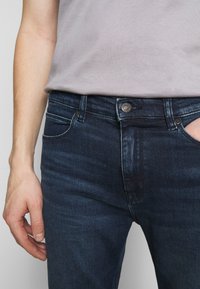 Tmavé denimové džíny se slim fit, s kovovou knoflíkem, předními kapsami a viditelnými švy. Materiál má hladkou strukturu a mírný lesk.