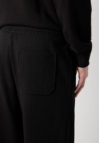 Zwarte sweatpants hebben een elastische tailleband en een rechthoekige achterzak. De stof lijkt zacht met een gladde textuur.